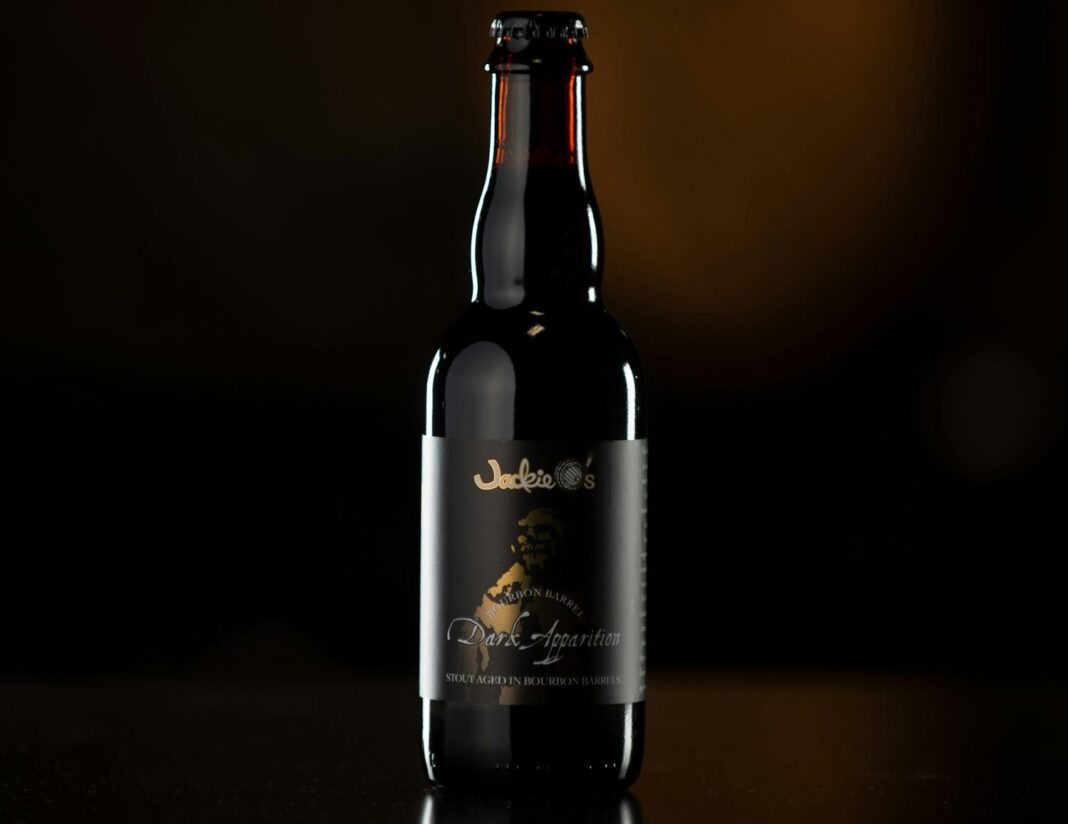 A bottle of Bourbon Barrel Dark Apparition with its distinctive label, set against a dark background, helps to highlight the image of this beer bottle.