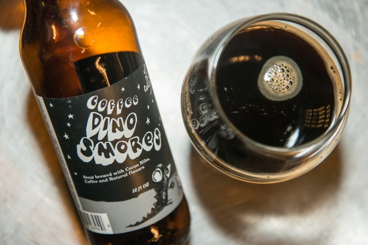 The image shows a glass of Dino Smores beer with a deep, dark black color - Dino S'mores: The Sweet Declaration of Off Color Brewing